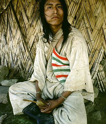 Colombia. Sierra Nevada de Santa Marta. Kogi Indian sits outside his straw hut. He is holding a gourd with a stick in it. He uses the stick to grind coca leaves, a mild narcotic,  and mix them with lime. One of his cheeks is inflated by a ball of that combination.